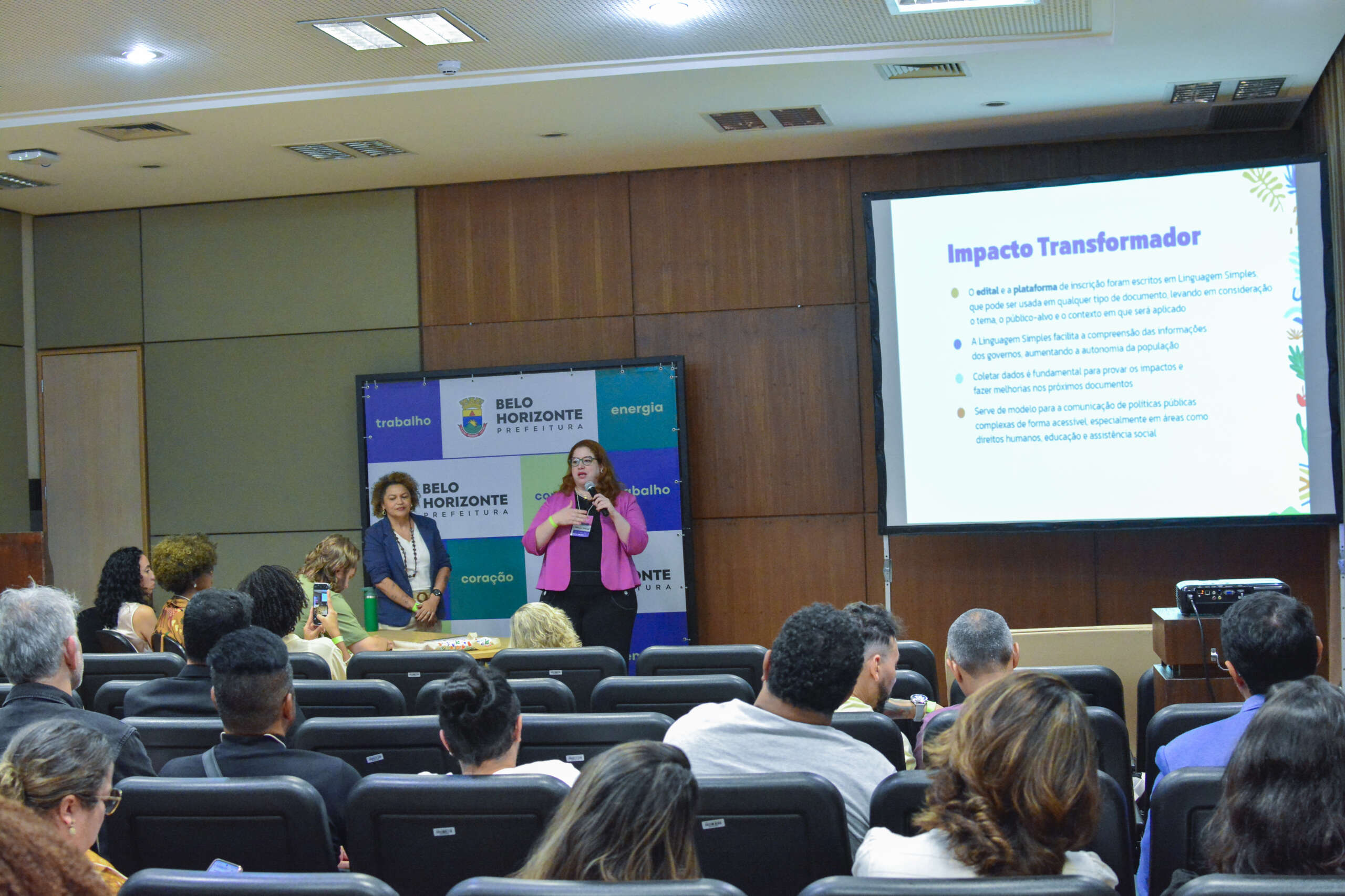 Uma mulher de cabelo escuro e camisa de manga longa dobrada nos cotovelos e calça jeans fala em um microfone no palco de um auditório, enquanto aponta para uma grande tela de apresentação à direita, que exibe o título "Impacto Transformador". Uma segunda mulher com cabelo escuro cacheado e blusa verde e preta está sentada à esquerda. Uma audiência diversa está sentada em cadeiras pretas de frente para o palco.