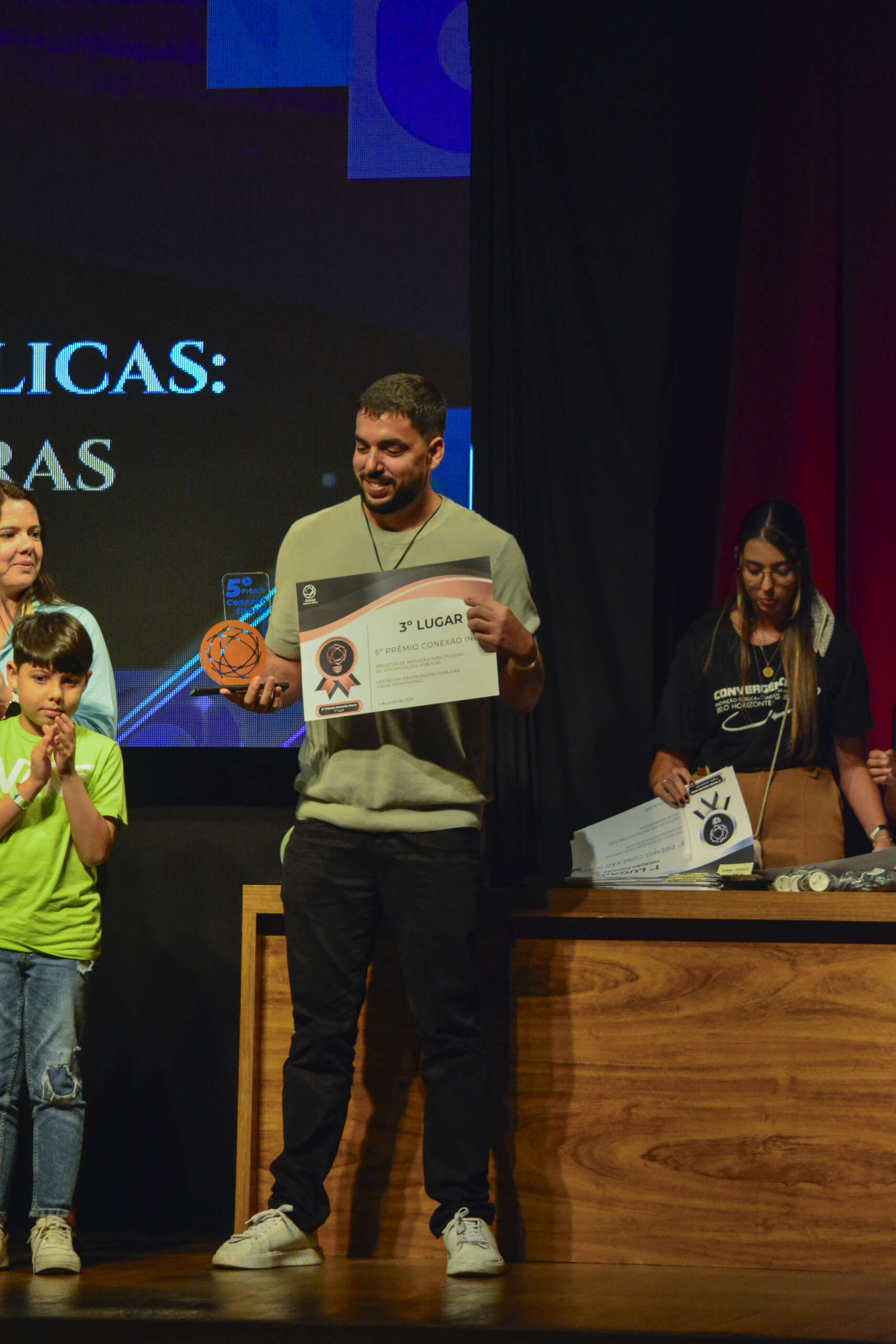 Um homem com barba, vestindo uma camiseta de algodão com elastico na parte de baixo, e calças escuras jeans, está no palco, sorrindo e segurando um certificado que diz "3º LUGAR". Ele também segura um pequeno troféu esférico laranja. À sua esquerda, uma mulher e uma criança de camiseta verde clara estão parcialmente visíveis. À sua direita, uma mulher com cabelo comprido está atrás de uma mesa de madeira, aparentemente organizando outros certificados.