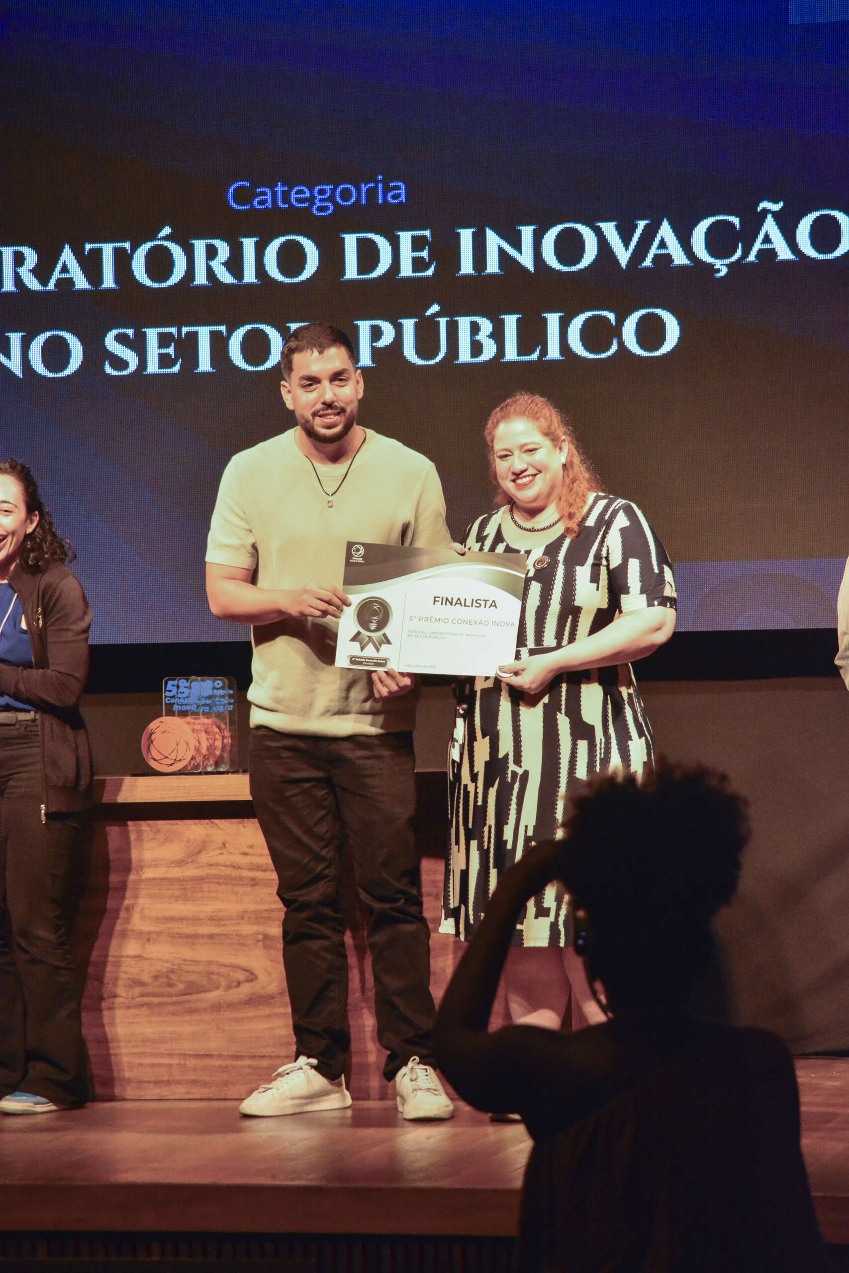 Um homem sorridente de camiseta verde-clara de algodão com elastico na parte de baixo e uma mulher sorridente com um vestido estampado em preto e branco estão no palco, segurando juntos um certificado que indica "FINALISTA". Atrás deles, uma tela de projeção exibe "Categoria" e parte do texto "LABORATÓRIO DE INOVAÇÃO NO SETOR PÚBLICO". Uma pessoa, em silhueta, pode ser vista em primeiro plano, na parte inferior direita da imagem, parecendo registrar o momento.