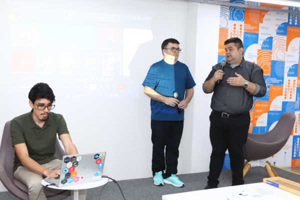 Três homens, dois em pé com microfones e um sentado com notebook, em sala de apresentação.