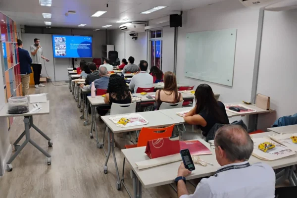 Homem de pé palestrando em sala de treinamento lotada com pessoas sentadas em mesas olhando a tela de projeção.