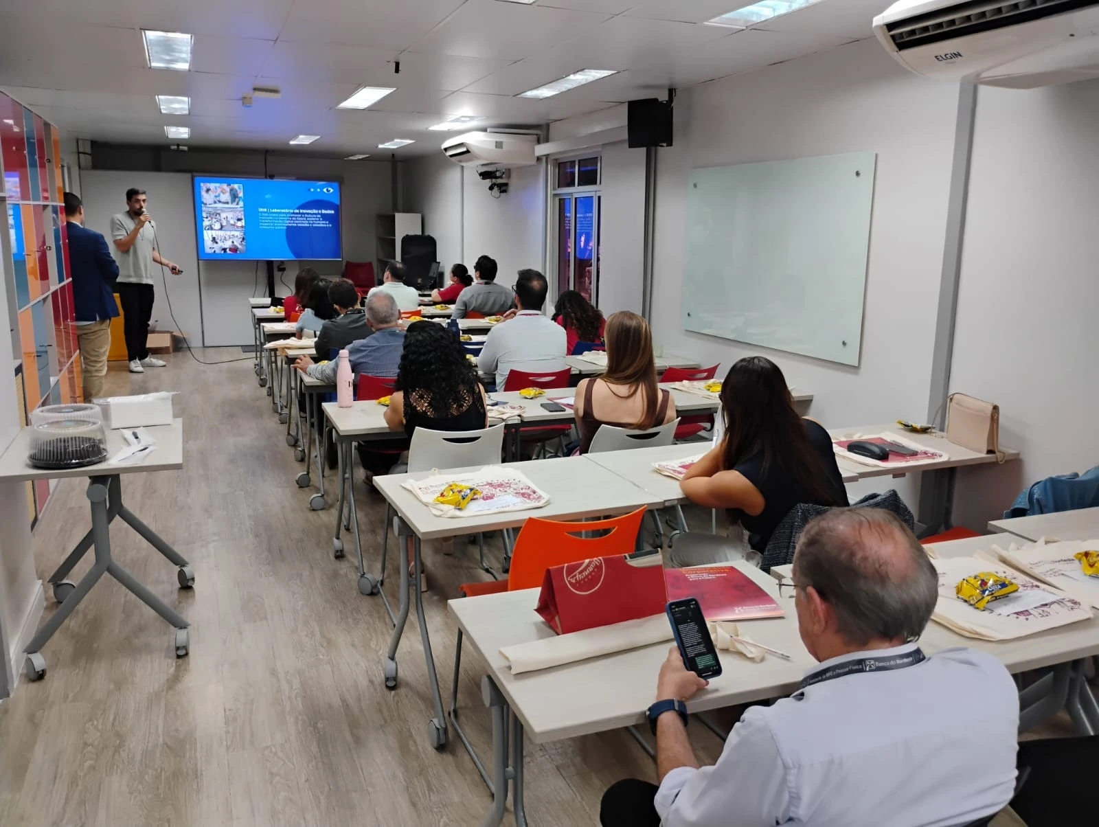 Homem de pé palestrando em sala de treinamento lotada com pessoas sentadas em mesas olhando a tela de projeção.