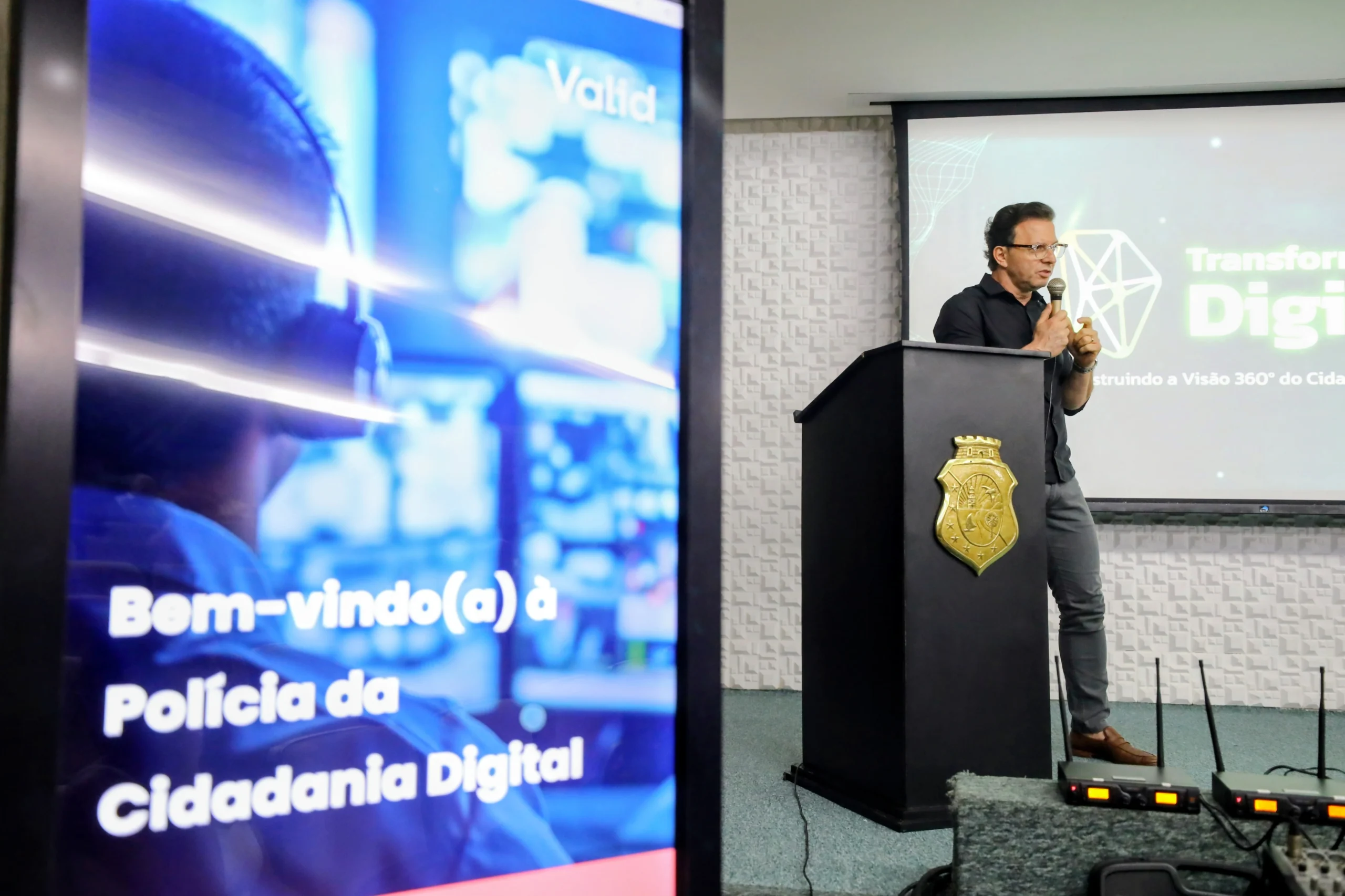 Acessor Vidal, realizando palestra, de camisa preta e óculos, falando ao microfone atrás de um púlpito com o brasão do governo do estado do ceará. O monitor lateral exibe a mensagem 'Bem-vindo(a) à Polícia da Cidadania Digital'.