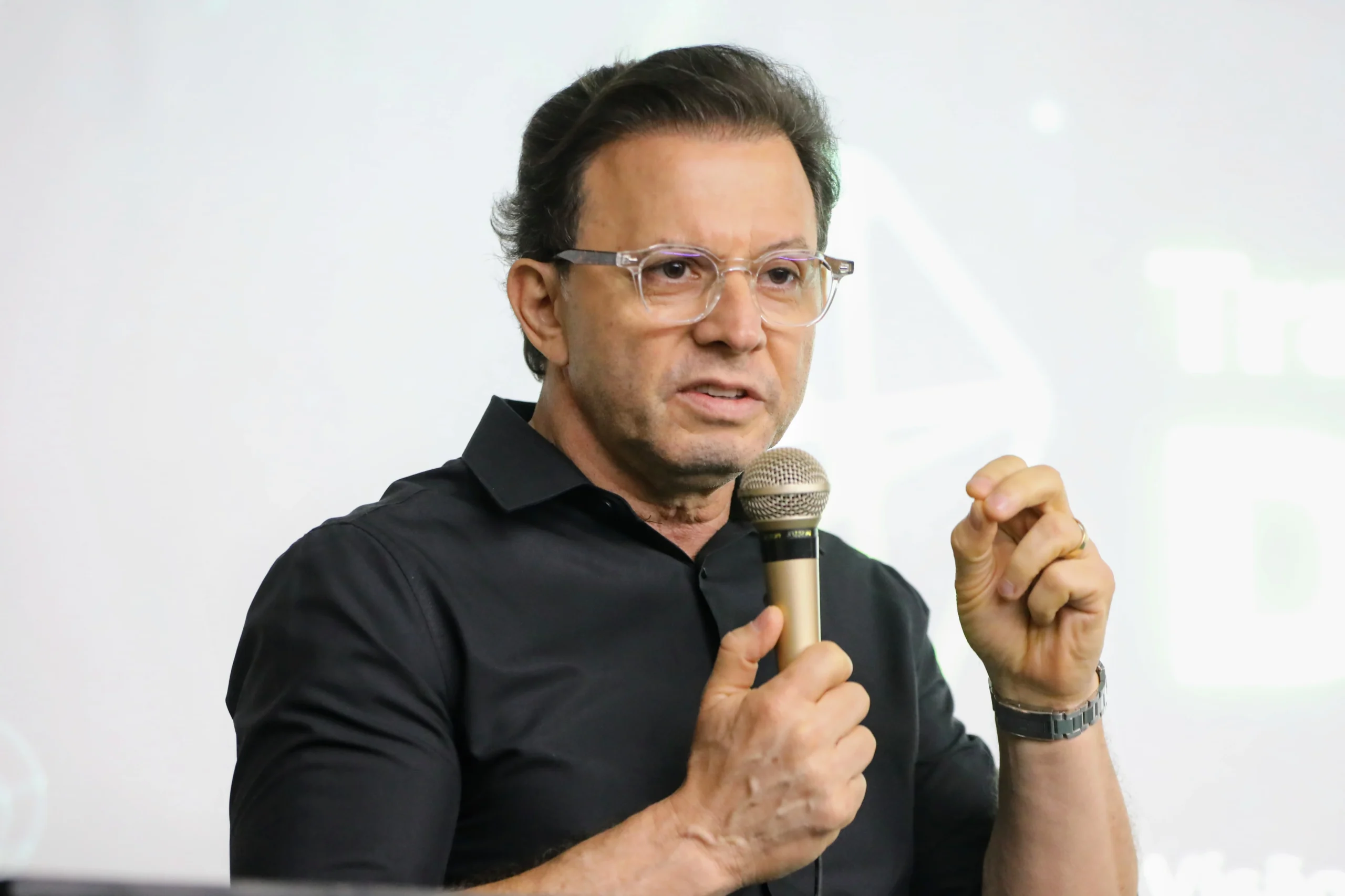 Close do acessor vidal, de óculos e camisa preta, falando em um microfone dourado e gesticulando com a mão direita.
