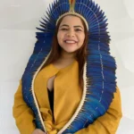 Mulher indígena sorridente de cabelos castanhos, vestindo um casaco amarelo e um grande cocar de penas azuis que desce pelos ombros. Ela está à frente de um fundo branco texturizado.