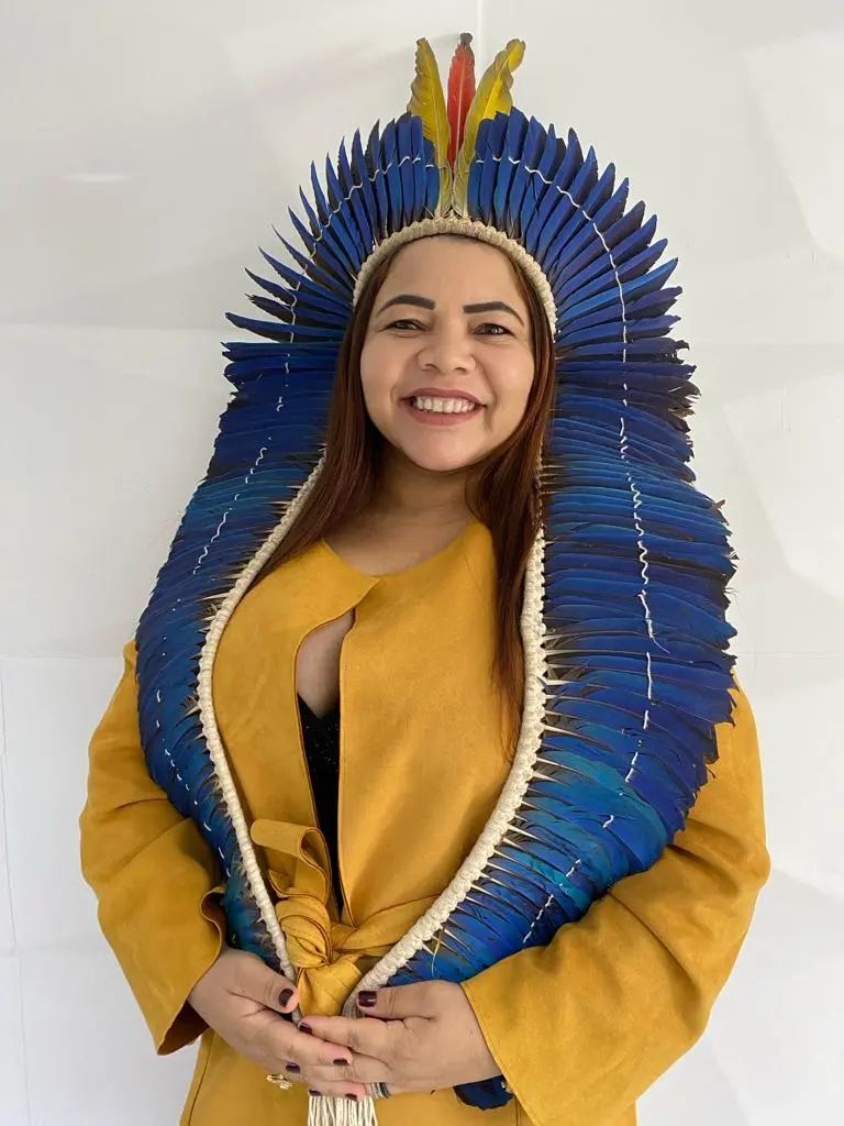 Mulher indígena sorridente de cabelos castanhos, vestindo um casaco amarelo e um grande cocar de penas azuis que desce pelos ombros. Ela está à frente de um fundo branco texturizado.